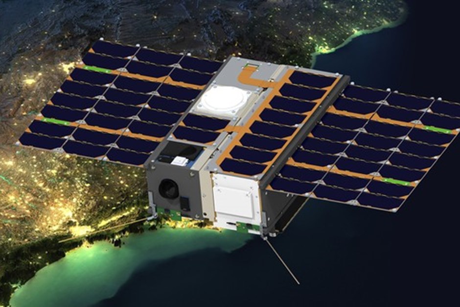 Uma foto do satélite VCUB1, formado por duas caixas estreitas unidas e várias placas de captação de energia solar. Ele voa acima do que parece ser a parte litorânea do Brasil à noite, com as luzes destacando os contornos do continente.